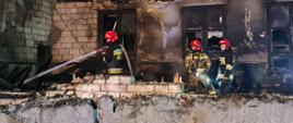Zdjęcie przestawia funkcjonariuszy Państwowej Straży Pożarnej oraz Ochotniczej Straży Pożarnej podczas działań gaśniczych. W tle pogorzelisko remizy - zniszczony obiekt. 