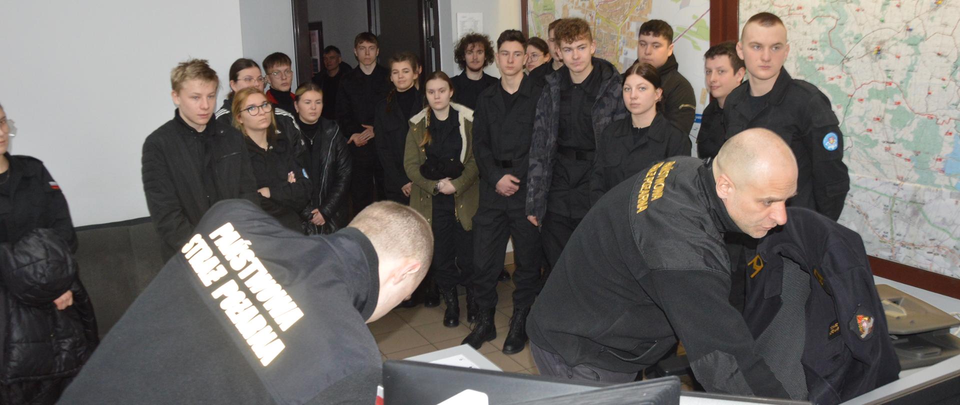 Wizyta klasy mundurowej. Stanowisko kierowania komendanta powiatowego PSP. Goście - uczniowie i uczennice ZS w Miejskiej Górce przyglądają się pracy dyżurnego. Prezentuje on odbiór "formatki" z WCPR. W tle mapy i drzwi. 