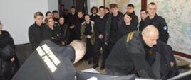 Wizyta klasy mundurowej. Stanowisko kierowania komendanta powiatowego PSP. Goście - uczniowie i uczennice ZS w Miejskiej Górce przyglądają się pracy dyżurnego. Prezentuje on odbiór "formatki" z WCPR. W tle mapy i drzwi. 