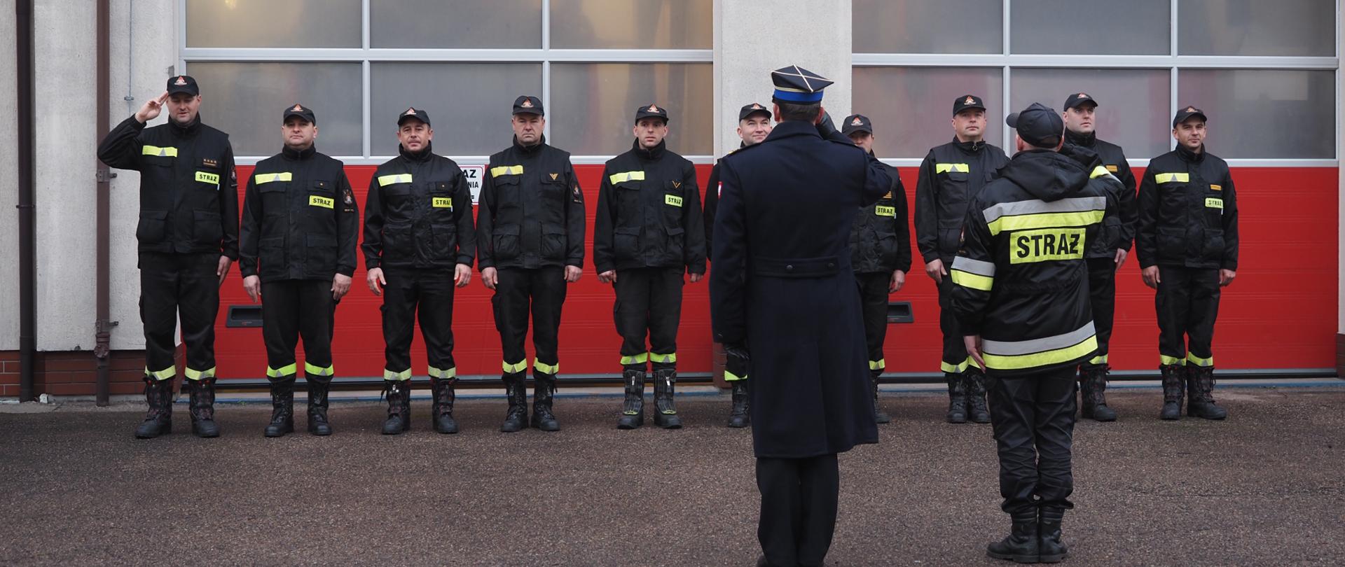 Strażacy ustawieni w szeregu przed budynkiem Komendy Powiatowej PSP w Bielsku Podlaskim podczas uroczystej zbiórki. Przed pododdziałem Zastępca Komendanta Powiatowego PSP w Bielsku Podlaskim bryg. Marcin Sawicki oraz Zastępca Dowódcy JRG kpt. Igor Strapczuk. Następuje wciągnięcie flagi państwowej na maszt i odśpiewanie Hymnu Narodowego.