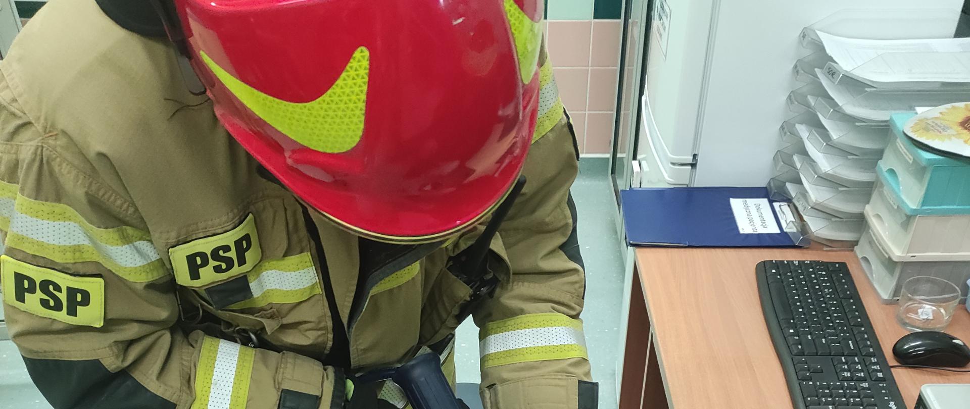 Pomieszczenie szpitala w Brzezinach, strażak w pełnym umundurowaniu bojowym przy pomocy narzędzi hydraulicznego w trakcie cięcia obrączki znajdującej się na opuchniętym palcu poszkodowanego mężczyzny.