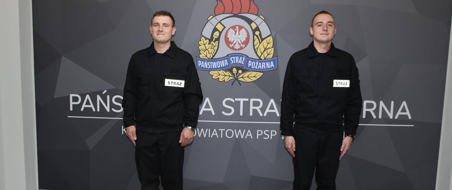 Zdjęcie przedstawia gabinet Komendanta Powiatowego Państwowej Straży Pożarnej w Łańcucie, na ścianie którego znajduje się tablica z logotypem Państwowej Straży Pożarnej. Na tle tablicy prezentują się nowo przyjęci strażacy.