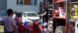 Zdjęcie przedstawia strażaka który stoi przy wozie strażackim i przedstawia sprzęt będący na wyposażeniu samochodu. Przed strażakiem stoi grupa dzieci wraz z nauczycielem i słuchają strażaka.