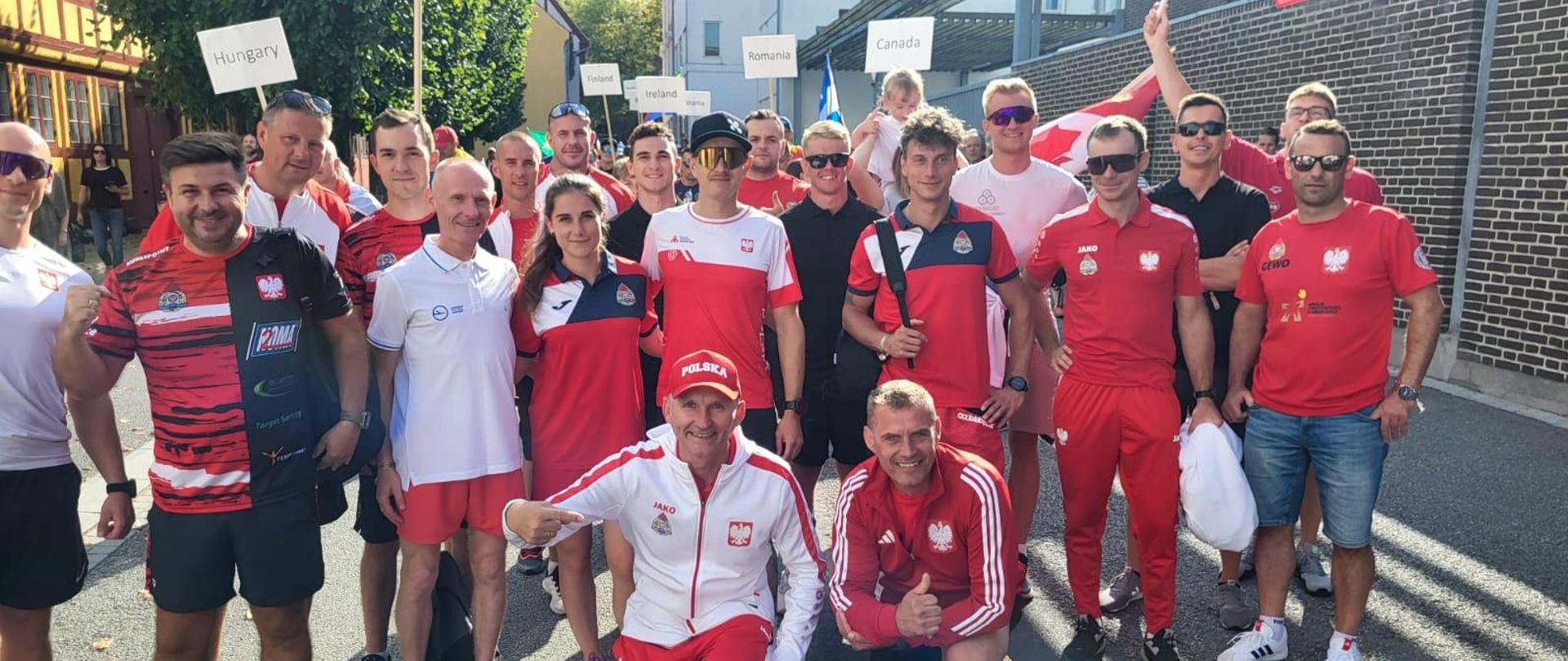 Na zdjęciu znajdują się strażacy reprezentujący Państwową Straż Pożarną w Światowych Igrzyskach Strażaków. Osoby znajdują się na ulicy, mają na sobie stroje sportowe. W tle widoczne są białe tabliczki z nazwami krajów.