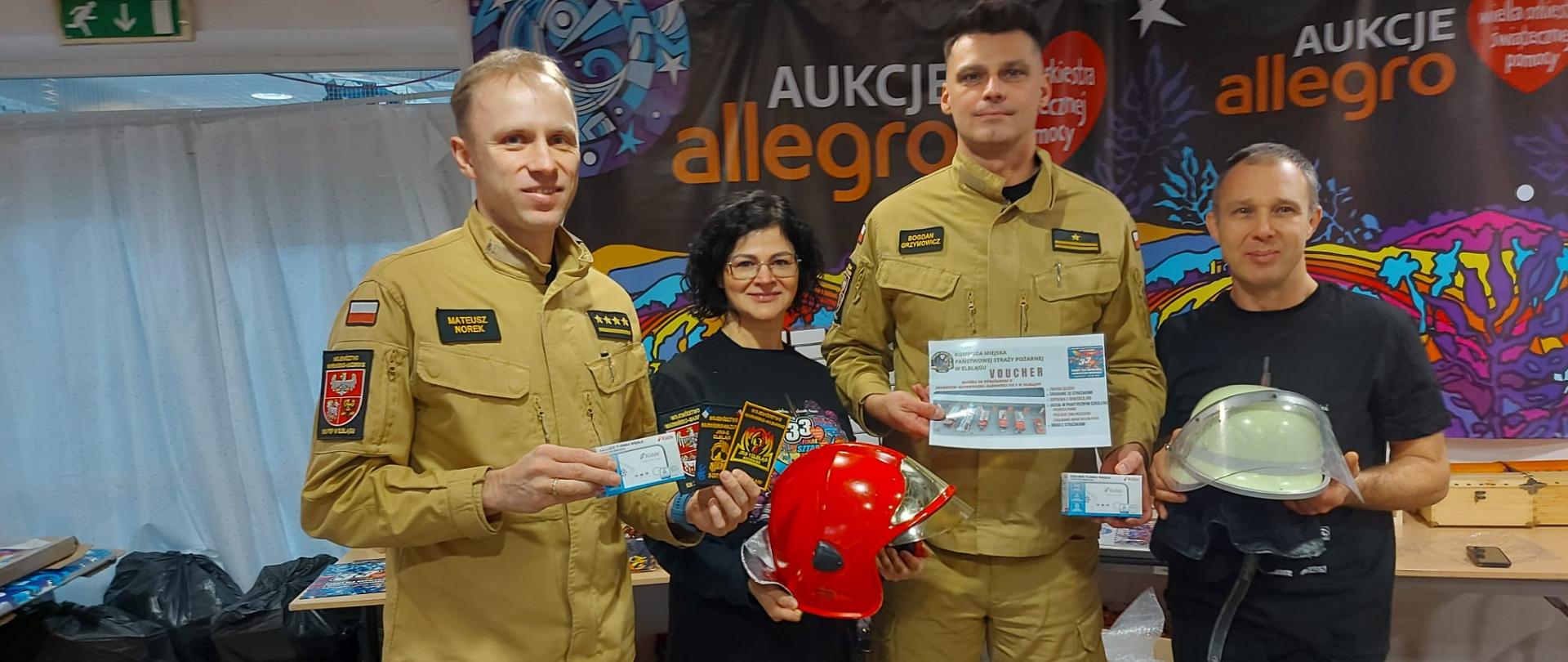 Funkcjonariusze Państwowej straży pożarnej prezentują w sztabie wielkiej orkiestry przedmioty przeznaczone na licytację, są to między innymi naszywki na mundury oraz czerwony hełm.
