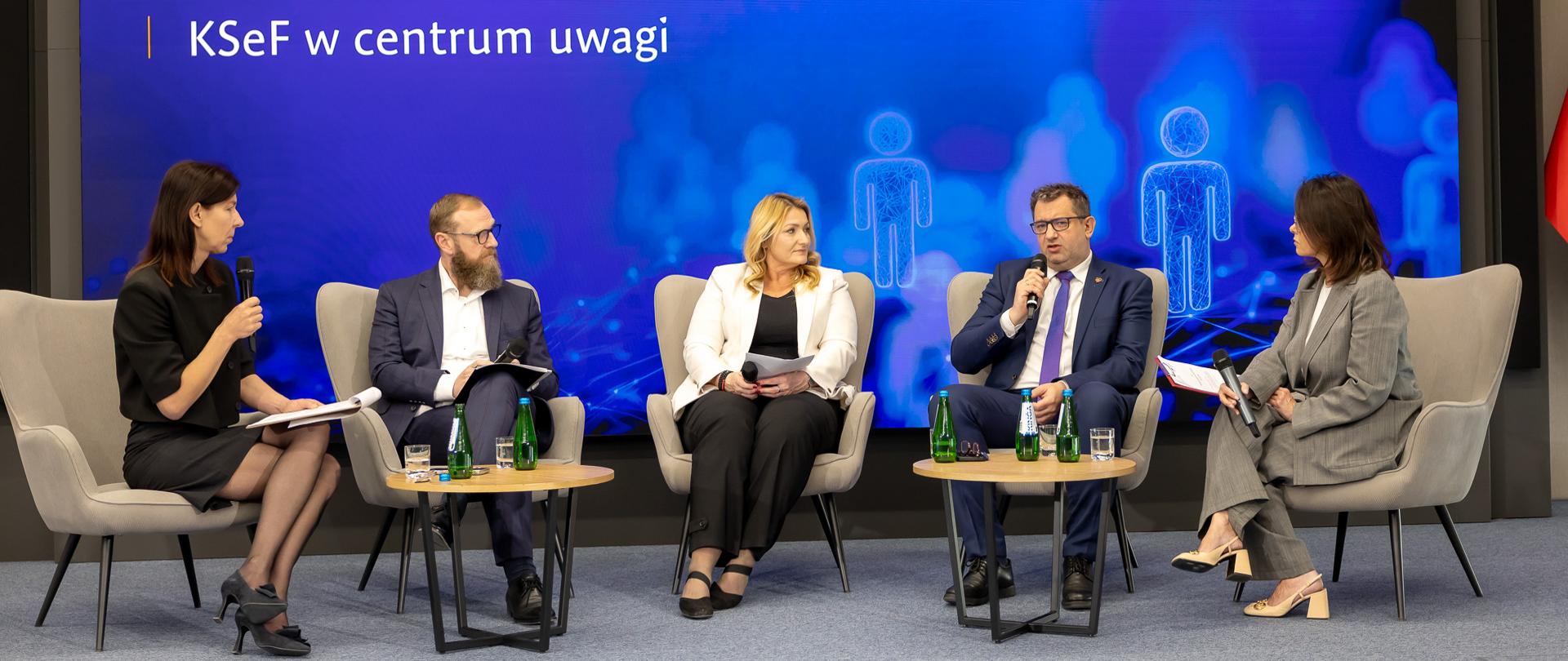 Marcin Łoboda Sekretarz Stanu w Ministerstwie Finansów przemawia do mikrofonu na tle ekranu informacyjnego wydarzenia.