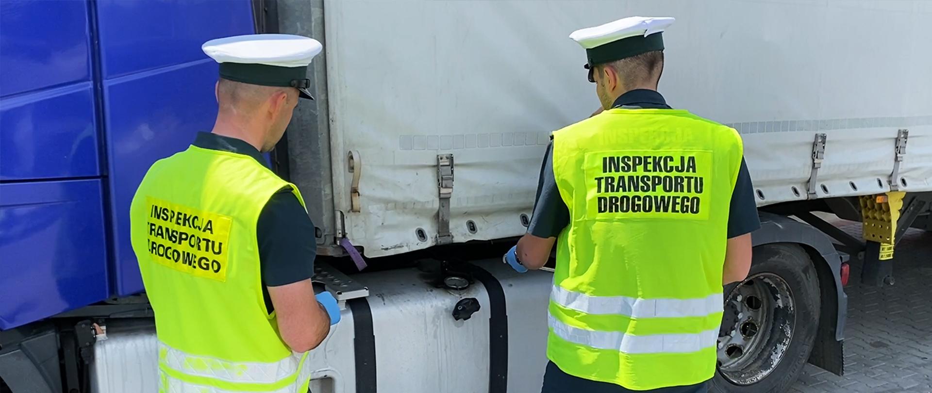 Insepktorzy w odblaskowych kamizelkach z napisem Inspekcja Transportu Drogowego stoją przy ciężarówce, bak od paliwa jest odkręcony, jeden z dwóch inspektorów trzyma w ręku wężyk, którym będzie pobierane paliwo do próbki

