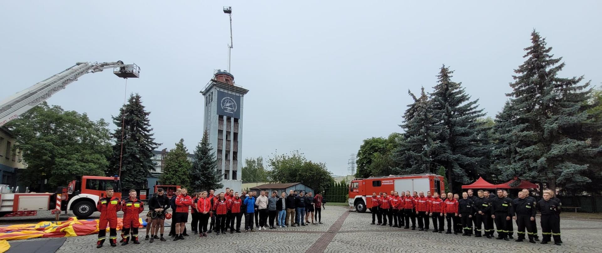 Na tle samochodu wysokościowego, wspinalni i drabiny mechanicznej stoją funkcjonariusze w dwóch grupach w dwuszeregu