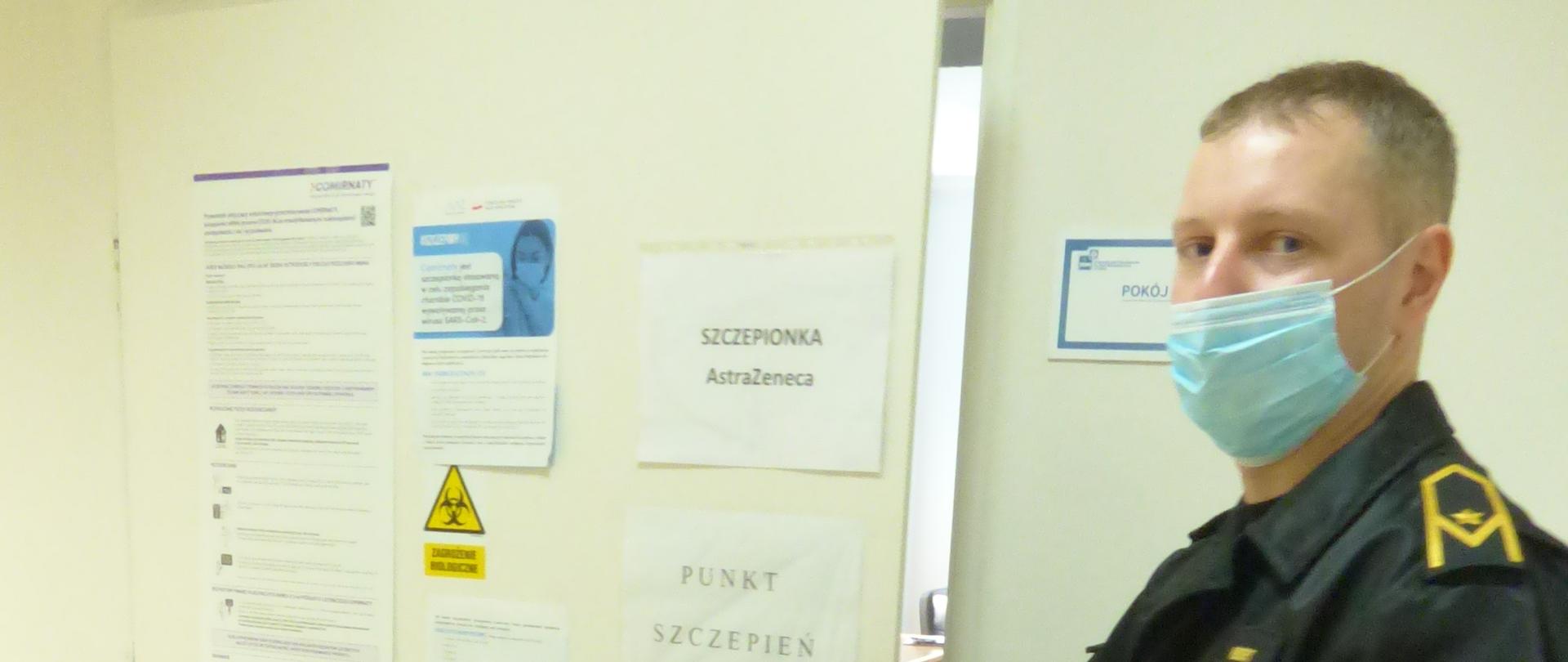 Strażak w maseczce chirurgicznej otwiera drzwi na których naklejona jest kartka z napisem punkt szczepień