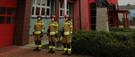 Na zdjęciu widzimy trzech strażaków stojących w szeregu ubranych w specjalne ubranie ochronne koloru piaskowego, na twarzach maski ochronne koloru białego. Na głowach czerwone hełmy z cyfrą "1". Za szeregiem strażaków uwidoczniona jest część masztu koloru białego. Dalej w kadrze widzimy część budynku komendy wraz z fragmentem pomnika strażaka trzymającego dziecko oraz roślinność dekoracyjną. Budynek komendy koloru łososiowego wraz z oszklonym wejściem głównym koloru czerwonego. Z lewej strony kadru widzimy część alarmowej bramy wyjazdowej koloru czerwonego. Z prawej strony kadru w oddali widzimy budynek mieszkalny ( kamienica dwukondygnacyjna ) koloru szarego oraz cześć banera reklamowego. 