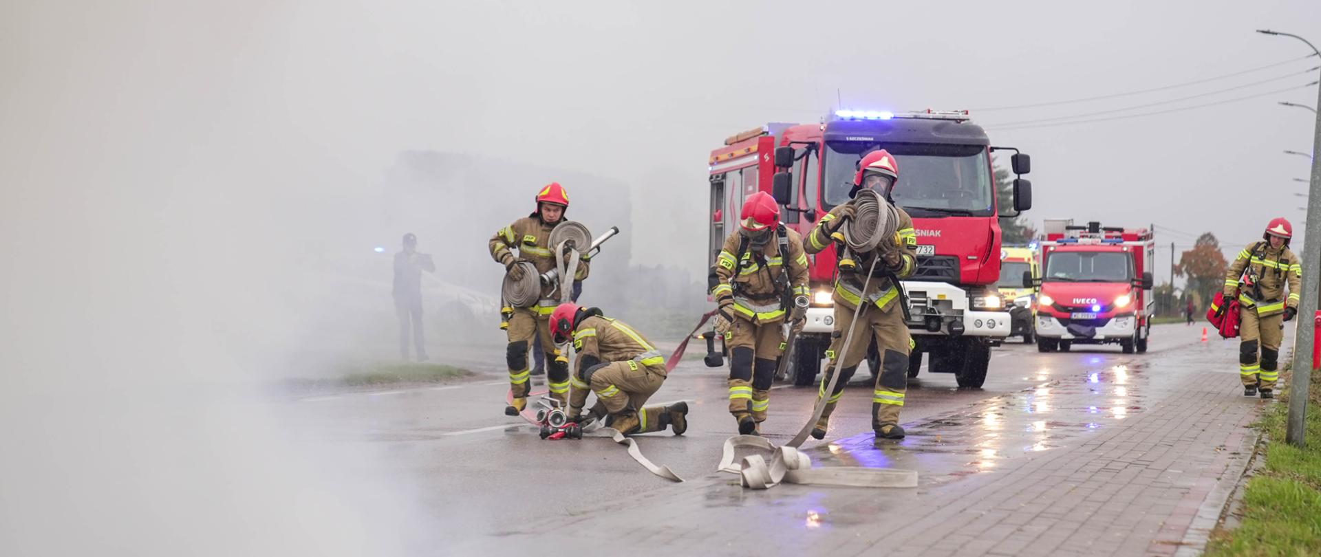 Strażacy PSP rozwijają węże i przygotowują sprzęt gaśniczy w zadymionym rejonie ulicy podczas ćwiczeń ratowniczych.