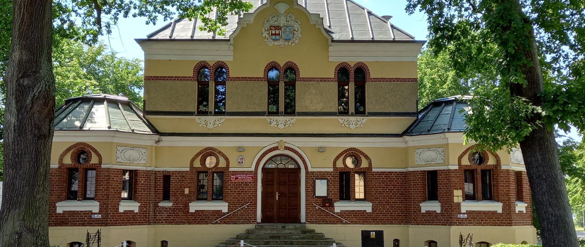Zdjęcie przedstawiające budynek szkoły muzycznej - elewacja frontowa. Budynek dwukondygnacyjny, symetryczny, z centralnym wejściem. Po obu stronach symetrycznie rozmieszczone baszty narożne, przed nimi na pierwszym planie dwa dęby. Elewacja częściowo z czerwonej cegły, częściowo tynkowana w kolorze żółtym.