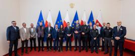 Grupa kilkunastu osób pozuje w jednym rzędzie wraz z Wojewodą Kujawsko-Pomorskim Michałem Sztyblem stojącym w centrum. Za uczestnikami ustawione są flagi Polski i Unii Europejskiej, a na ścianie wisi godło. Część osób ma na piersiach przypięte pamiątkowe medale. 