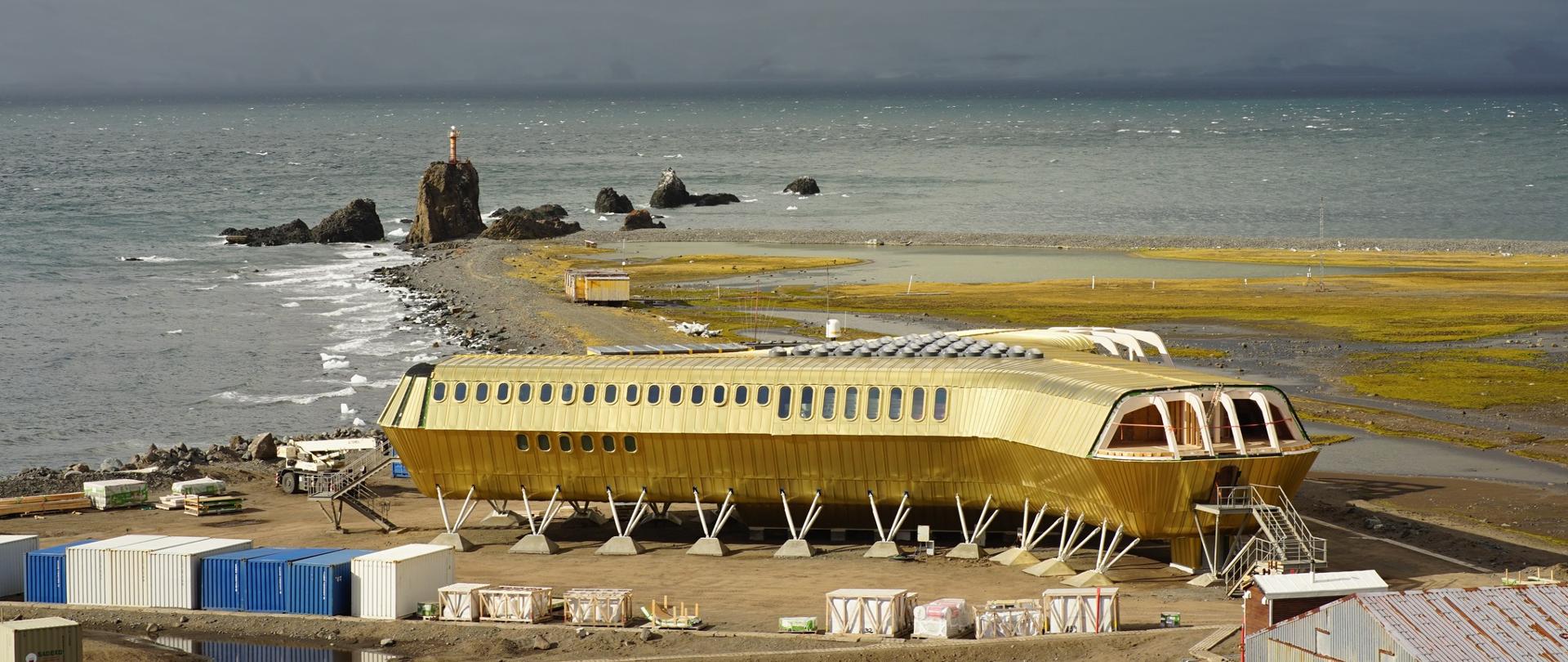 Na tle szarego morza, na szarożółtym terenie stoi złocisty budynek w kształcie trójłopatowego śmigła.
