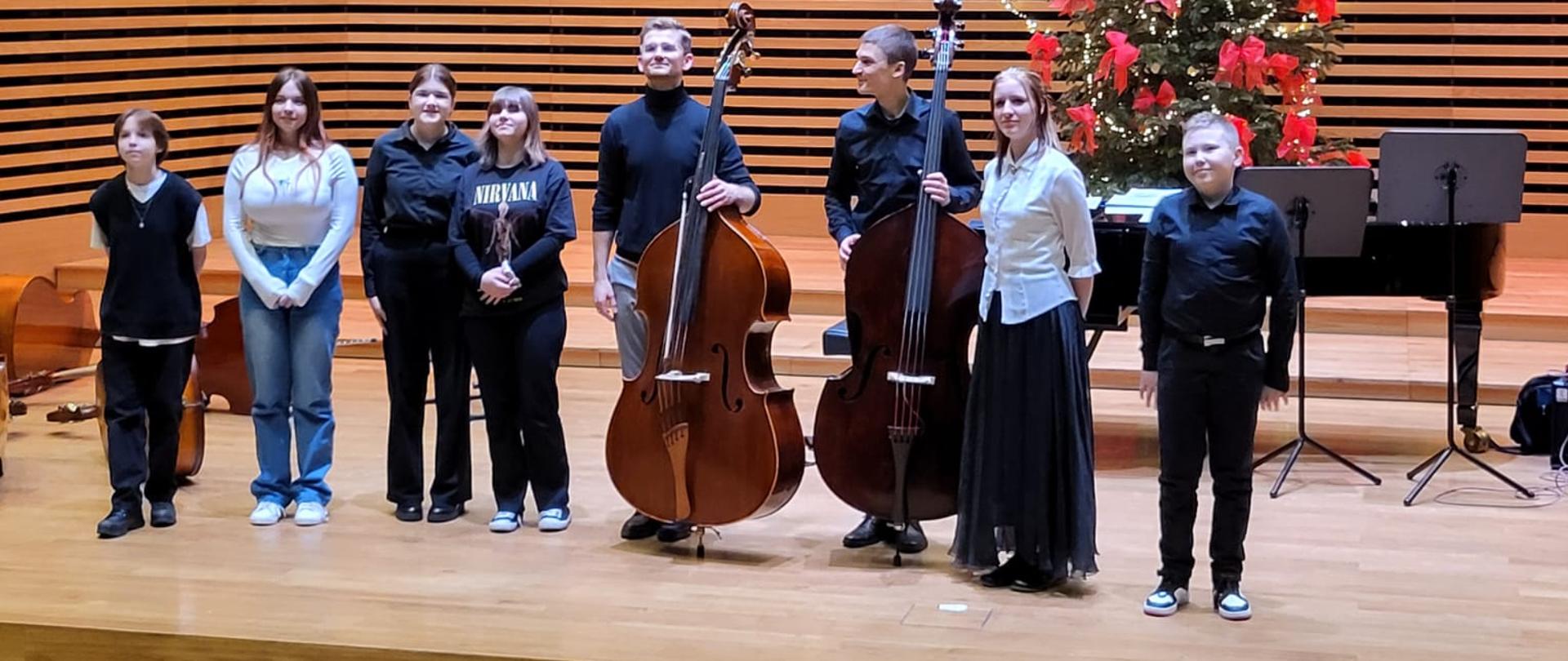 Dwóch mężczyzn trzymających kontrabasy oraz pięć dziewcząt i jeden chłopiec stoją w jednym rzędzie na estradzie sali koncertowej.
