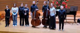 Dwóch mężczyzn trzymających kontrabasy oraz pięć dziewcząt i jeden chłopiec stoją w jednym rzędzie na estradzie sali koncertowej.