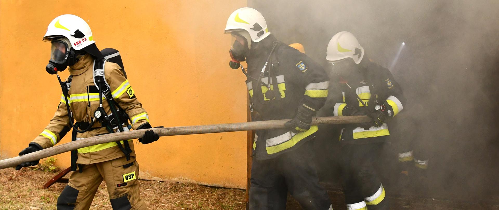 czterech strażaków ubranych w sprzęt ODO wychodzi z nawodniona linia z pomieszczenia z którego wydobywa się dym