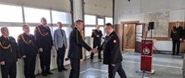 Pomorski komendant wojewódzki Państwowej Straży Pożarnej gratuluje funkcjonariuszowi podając mu dłoń. Obok stoją zaproszeni goście.