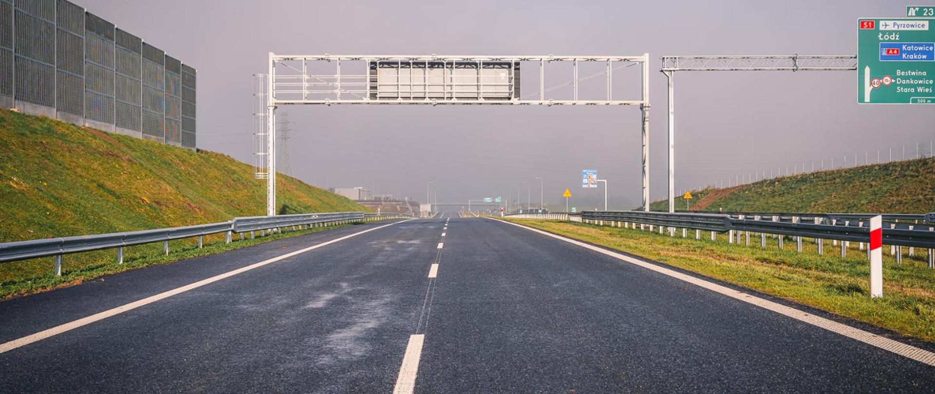 Nowo wybudowany odcinek drogi ekspresowej, widok na dwupasmową jezdnię z barierami energochłonnymi, ekranami akustycznymi po lewej stronie oraz bramownicą nad jezdnią; w tle znaki kierunkowe i lekkie zamglenie.