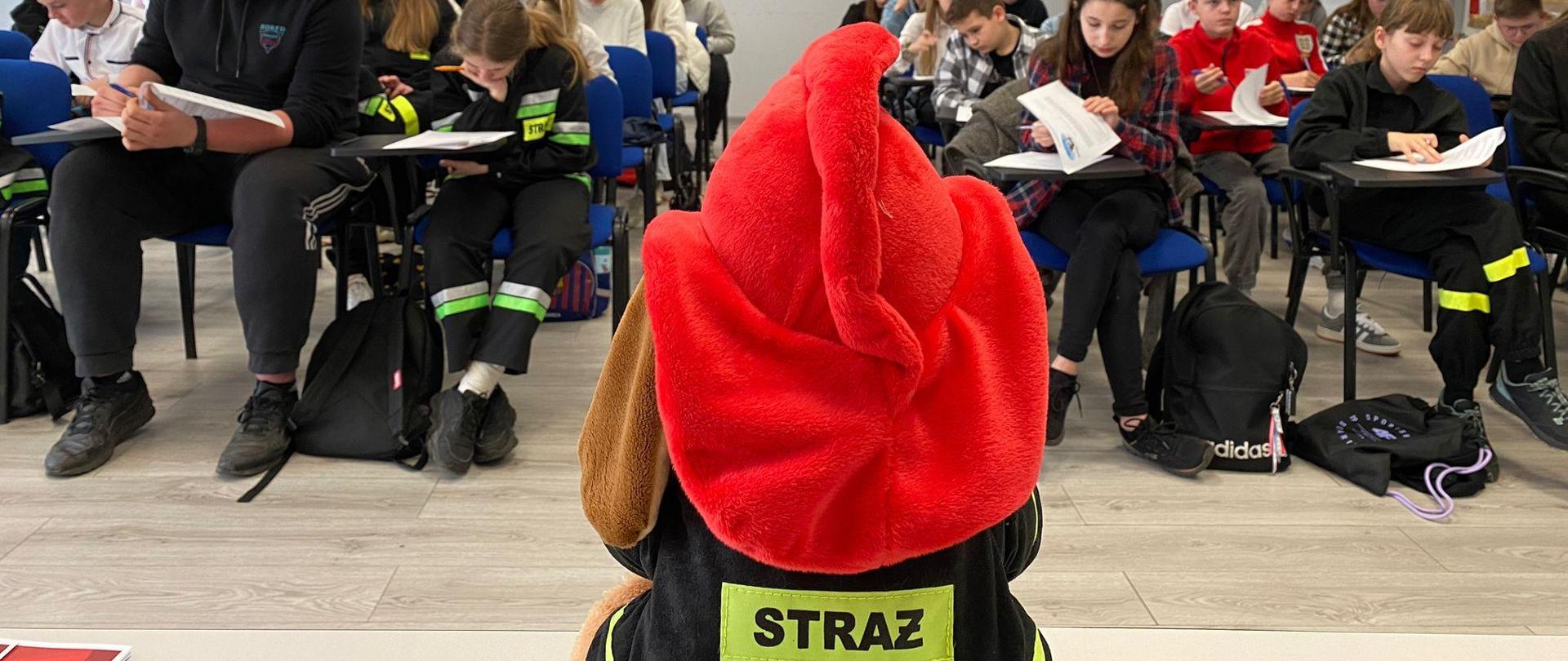 Zdjęcie wykonane wewnątrz pomieszczenia. Na pierwszym planie maskotka z napisem STRAŻ. Na drugim planie uczestnicy turnieju wraz z opiekunami siedzą na krzesłach przygotowani do testu. 