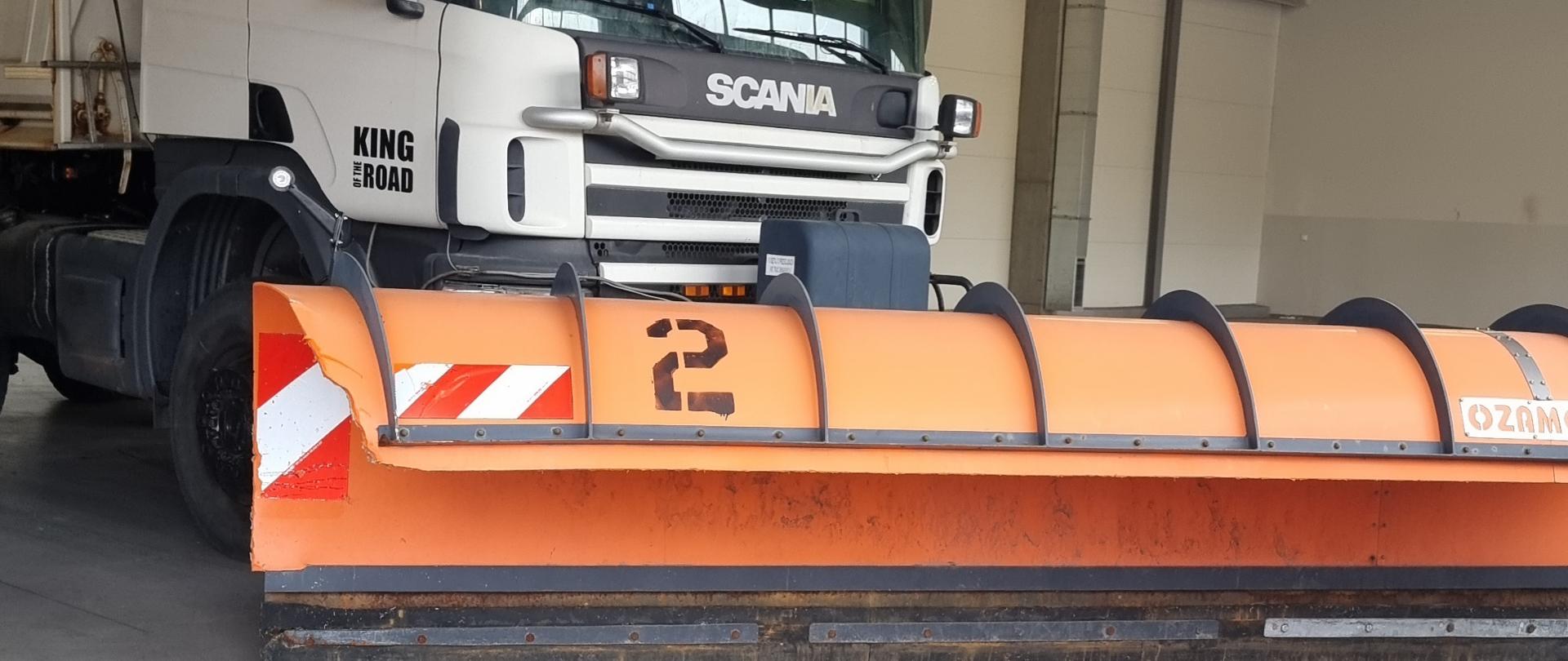 Na zdjęciu znajduje się biały samochód ciężarowy marki Scania, ustawiony w dużym, dobrze oświetlonym garażu lub hali serwisowej. Pojazd jest wyposażony w masywny pomarańczowy pług śnieżny z odblaskowymi biało-czerwonymi pasami, zamontowany na przedniej części. W tle widoczne są metalowe konstrukcje, przewody i lampy sufitowe, typowe dla obiektów technicznych. Podłoga jest gładka, w kolorze szarym, a całość sprawia wrażenie przygotowań do sezonu zimowego.