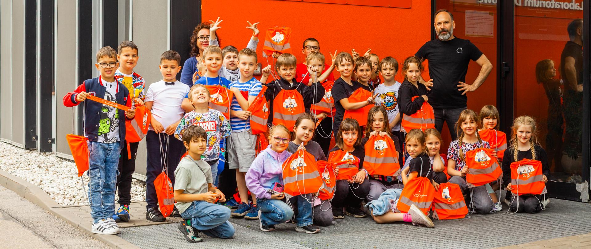 Na zdjęciu widoczna jest grupa dzieci stojących przed budynkiem oznaczonym napisem: „GDDKiA Oddział w Katowicach Wydział Technologii i Jakości Budowy Dróg Laboratorium Drogowe”. Dzieci ubrane są w pomarańczowe koszulki i trzymają pomarańczowe torby, co może sugerować udział w zorganizowanej wycieczce lub wydarzeniu edukacyjnym. Obok grupy stoi dorosły mężczyzna ubrany na czarno, prawdopodobnie opiekun lub przewodnik.