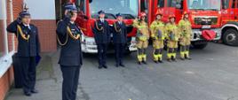 Strażacy stoją podczas zbiórki z okazji Święta Niepodległości. W tle stoją samochody pożarnicze.