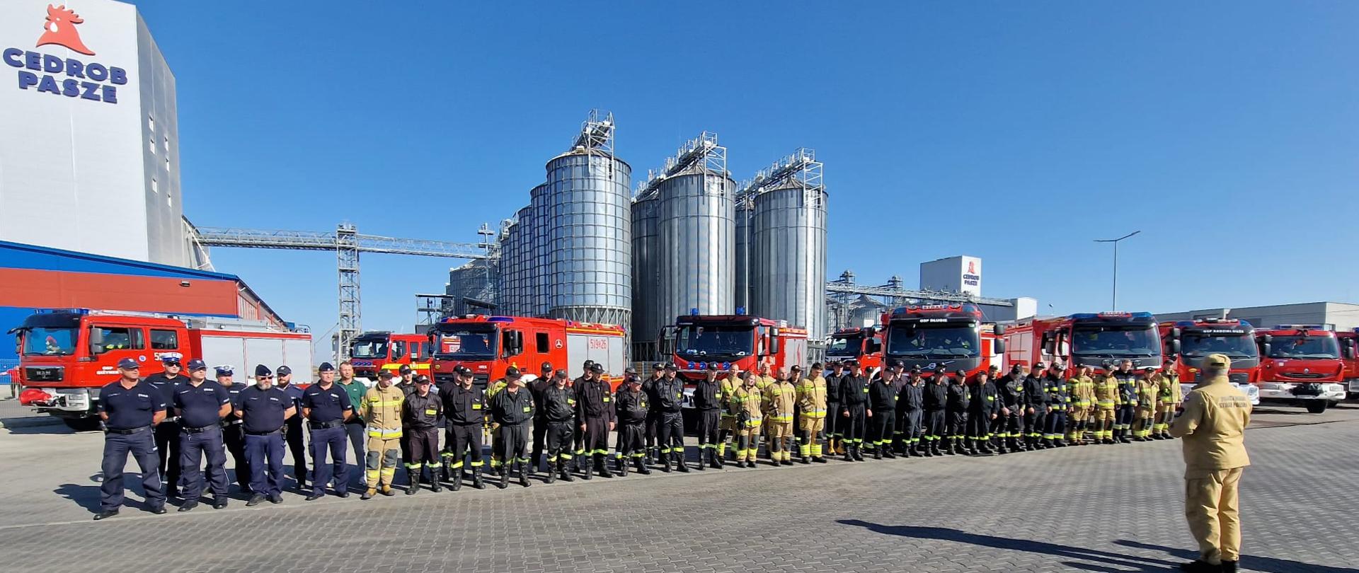Na zdjęciu widzimy zbiórkę strażaków biorących udział w ćwiczeniach CEDROB 20023 na tle zakładu produkcyjnego Cedrob S.A.