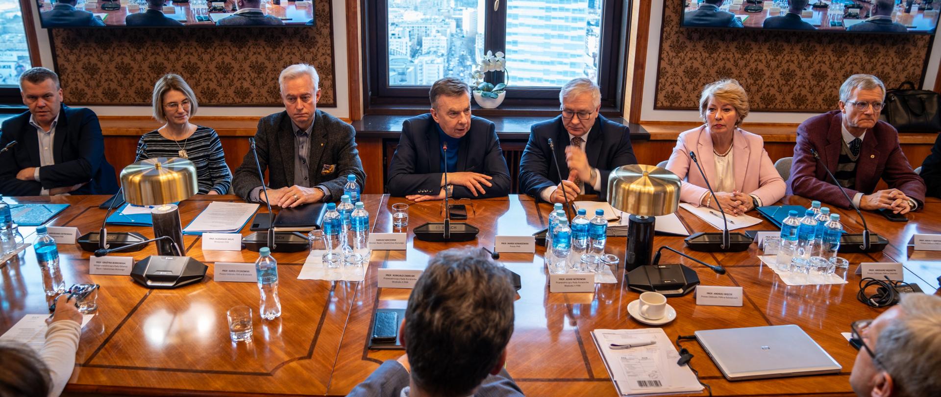 Zdjęcie z góry, za drewnianym prostokątnym stołem siedzi minister Wieczorek i kilkanaście innych osób.