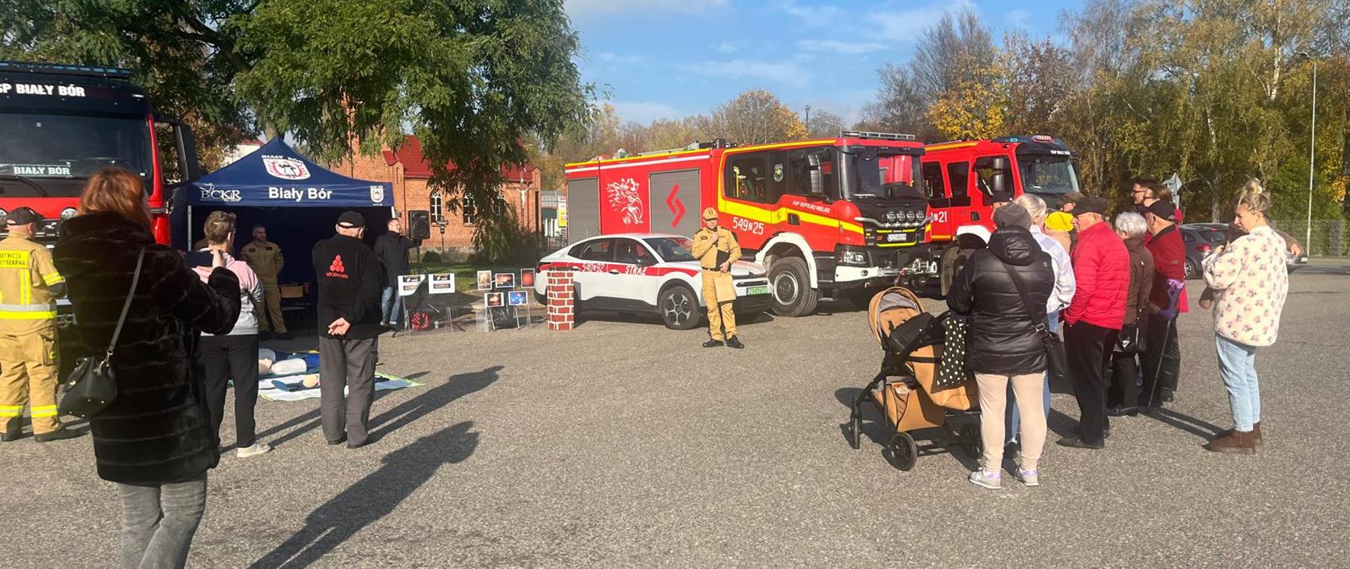 Na dużym parkingu odbywa się pokaz strażacki w ramach programu „Czujne Zachodniopomorskie”, Festyn Bezpieczeństwa. Grupa ludzi stoi i obserwuje strażaków prezentujących sprzęt i manekiny do treningu resuscytacji obok czerwonych wozów strażackich. Po lewej stronie znajduje się niebieski namiot z napisem „Biały Bór”. Widoczne są także małe dzieci, w tym jedno w wózku. W tle drzewa pokryte jesiennymi liśćmi i budynki. Słoneczny dzień rzuca wyraźne cienie uczestników wydarzenia na asfalt.