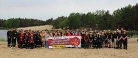 zdjęcie przedstawia nłodych druchów i druchny, prowadzących oraz zaproszonych gości na plaży z plakatem o napisie "zjazd młodzieżowych drużyn pożarniczych" w tle zalew oraz drzewa
