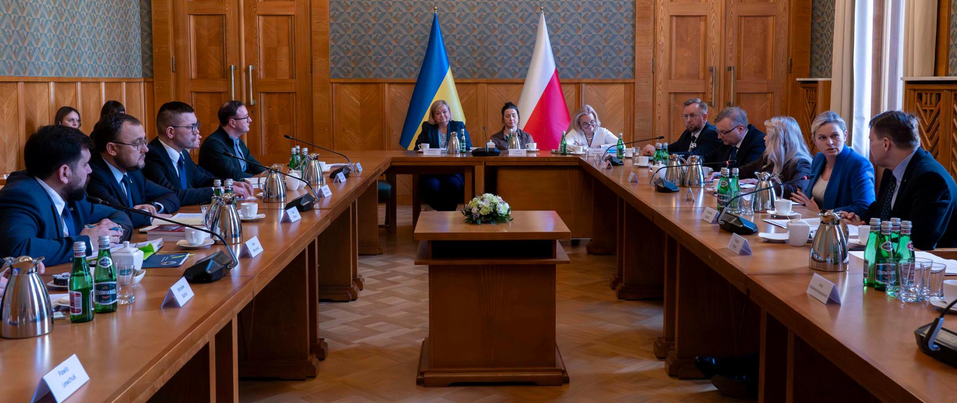 Szerokie ujęcie sali konferencyjnej. Przy długim stole po prawej stronie siedzą m.in. Barbara Nowacka i Radosław Sikorski. Minister Nowacka gestykuluje podczas rozmowy z siedzącymi naprzeciwko przedstawicielami Ukrainy. W tle, na środku ściany, widoczne są flagi Ukrainy i Polski.