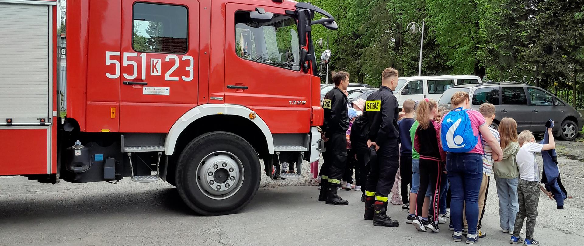 Widok z tyłu. Dzieci i strażacy stoją przed samochodem strażackim i pozują do pamiątkowej fotografii. 