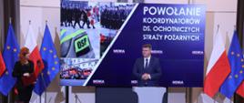 Sekretarz stanu w Ministerstwie Spraw Wewnętrznych i Administracji przemawia obok stoi kobieta a za nimi stoją flagi Unii Europejskiej oraz Polski telebim na którym wyświetlany jest napis powołanie koordynatorów ds. ochotniczych straży pożarnych.