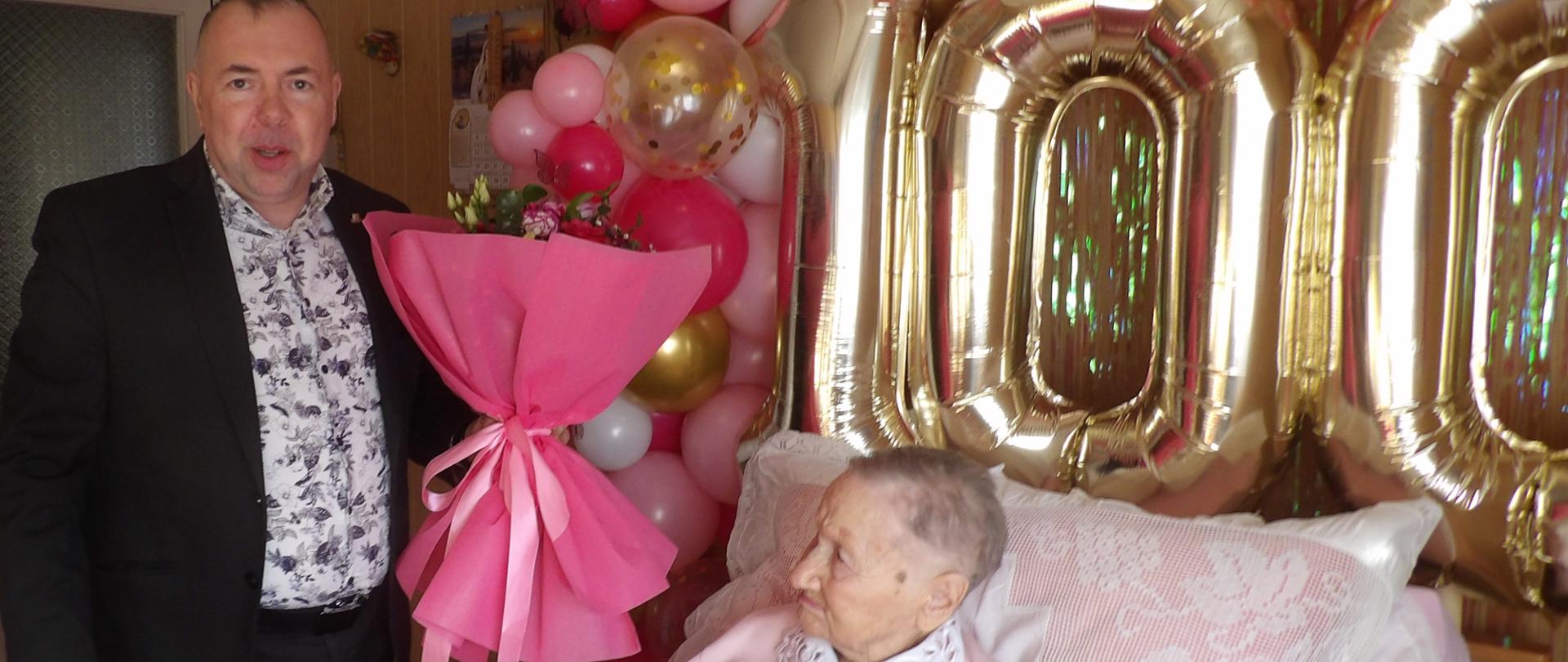 Zdjęcie przedstawia uroczystość 100. urodzin starszej kobiety w domowej atmosferze. Jubilatka, ubrana w różowy sweter z koronkowym kołnierzykiem, odpoczywa w łóżku rehabilitacyjnym, trzymając bukiet czerwonych róż. Obok niej stoi uśmiechnięty mężczyzna w marynarce z drugim, dużym bukietem. Tło zdobi girlanda z biało-różowo-złotych balonów oraz wielkie, złote cyfry tworzące liczbę 100. Scena emanuje ciepłem i szacunkiem dla wyjątkowego jubileuszu.