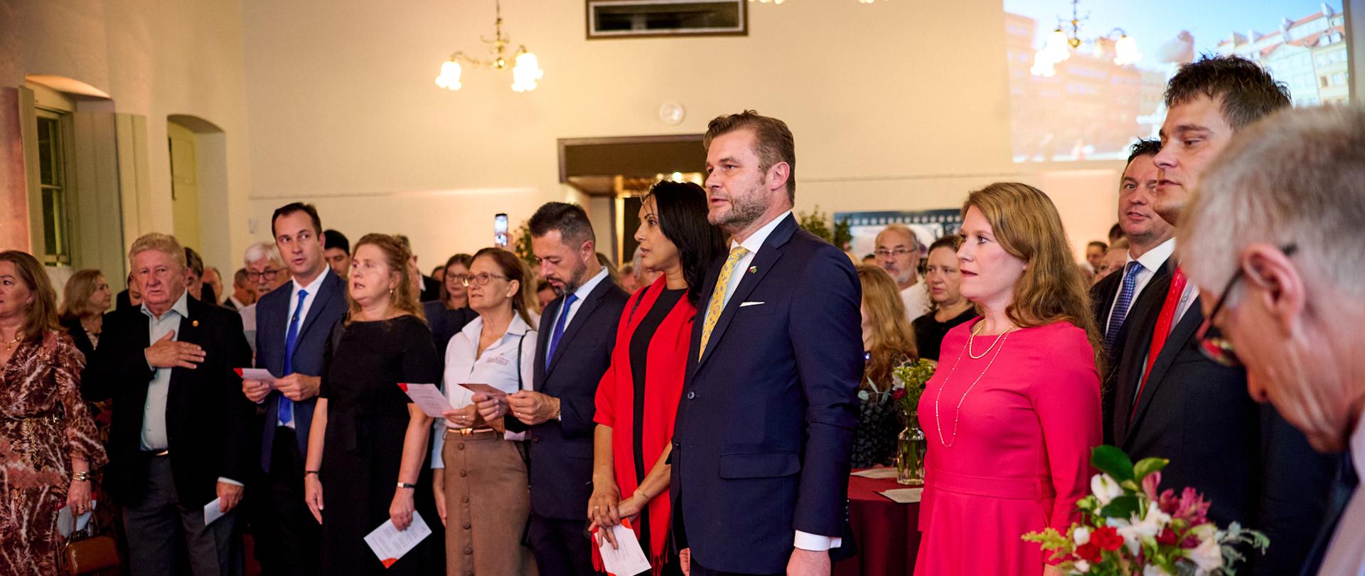 W historycznym wnętrzu, nastrojowo podświetlonym i z dekoracjami z biało-czerwonych kwiatów, widać wiele osób stojących w półkolu, na baczność, uroczyście ubranych i w podniosłym poważnym nastroju śpiewających hymn. 