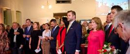 W historycznym wnętrzu, nastrojowo podświetlonym i z dekoracjami z biało-czerwonych kwiatów, widać wiele osób stojących w półkolu, na baczność, uroczyście ubranych i w podniosłym poważnym nastroju śpiewających hymn. 
