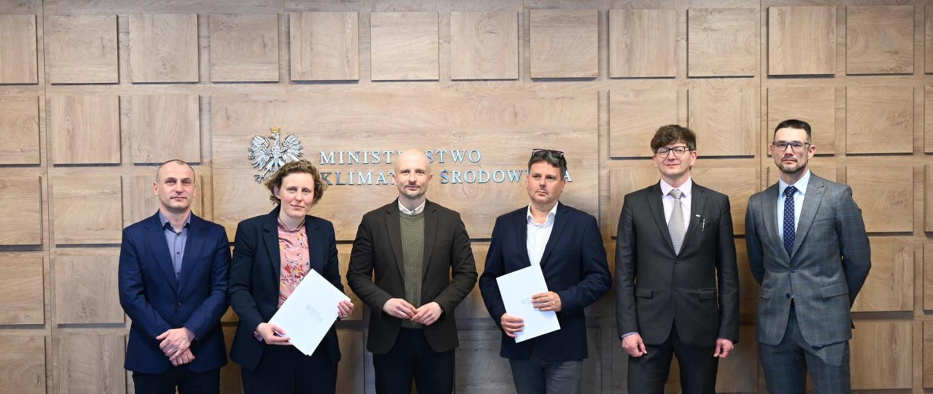 A meeting regarding the selection of the contractor for the design documentation of the Central Animal Shelter. The photo shows five people, including Deputy Minister of Climate and Environment Mikołaj Dorożała, Director of the Central Animal Shelter Agnieszka Czujkowska, DVM, and the contractor’s representative Tomasz Kacprowicz. All of them are smiling, and behind them the words “Ministry of Climate and Environment” can be seen.