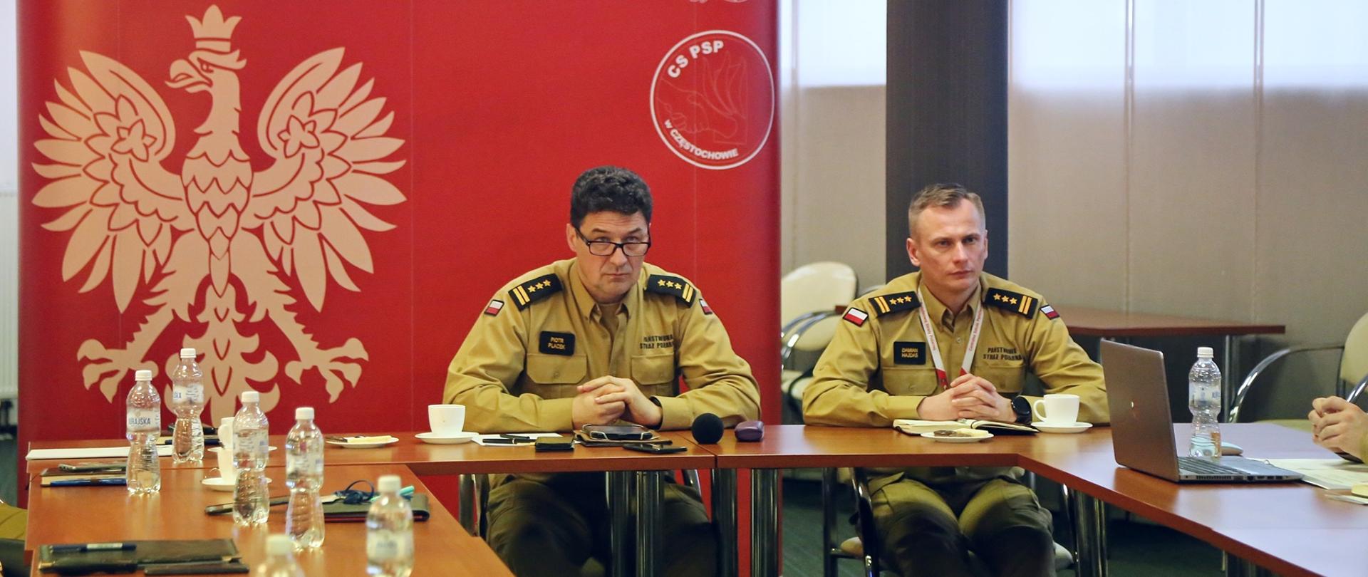 Komendant Centralnej Szkoły PSP wraz ze swoim Zastępcą siedzą za stołem podczas narady służbowej, w tle baner reklamowy CS PSP