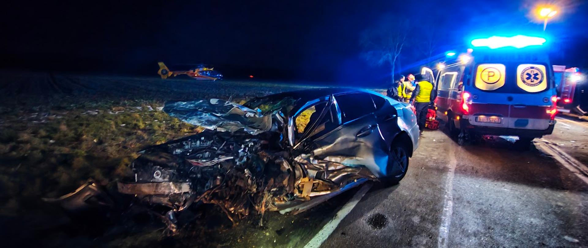 Zdjęcie przedstawia samochód osobowy po czołowym zderzeniu z innym pojazdem, w tle śmigłowiec Lotniczego Pogotowia Ratunkowego oraz karetka 