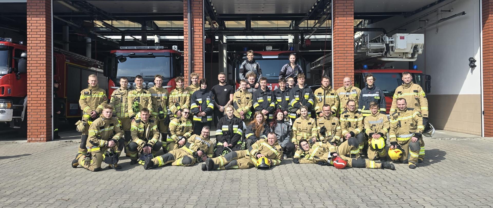 Zdjęcie przedstawia dużą grupę strażaków pozujących do wspólnej fotografii przed budynkiem remizy. Jest to jasny, słoneczny dzień.
Otoczenie i wyposażenie
• Budynek: W tle widać nowoczesną remizę strażacką z kilkoma stanowiskami garażowymi. Bramy są otwarte, odsłaniając zaparkowane wewnątrz wozy strażackie.
• Pojazdy: W garażach widoczne są co najmniej trzy czerwone samochody pożarnicze, w tym jeden wyposażony w drabinę mechaniczną lub podnośnik.
• Podłoże: Grupa stoi i leży na placu wyłożonym szarą kostką brukową.
Osoby na zdjęciu
Na fotografii znajduje się kilkadziesiąt osób (około 30-40). Większość z nich jest ubrana w profesjonalne, piaskowe (beżowe) mundury bojowe, typowe dla nowoczesnych jednostek straży pożarnej.
• Ubiór: Niektórzy strażacy mają na sobie ciemniejsze (granatowe lub czarne) ubrania służbowe lub koszulki. Część osób trzyma w dłoniach lub ma położone przed sobą hełmy strażackie (widoczne kolory to żółty, biały i czerwony).
• Ustawienie: Grupa jest ustawiona wielopoziomowo:
o Osoby w tylnych rzędach stoją.
o Kilka osób siedzi lub klęczy w środkowej części.
o Trzy osoby leżą na boku w swobodnych pozach na samym przodzie grupy