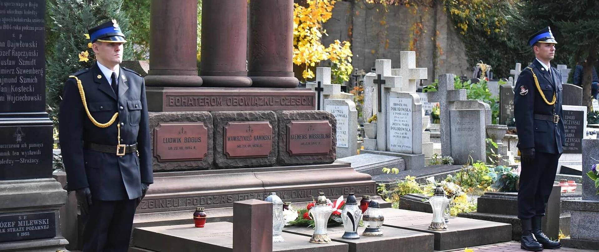 na zdjęciu dwóch strażaków w umundurowaniu reprezentacyjnym stoi jako warta honorowa przed pomnikami poległych w akcjach strażaków. Pomnik po lewej jest w formie monolitu w kolorze czarnym drugi trzykolumnowy z czerwonego bazaltu, na nagrobkach napisy z imionami i nazwiskami, w tle inne nagrobki i drzewa