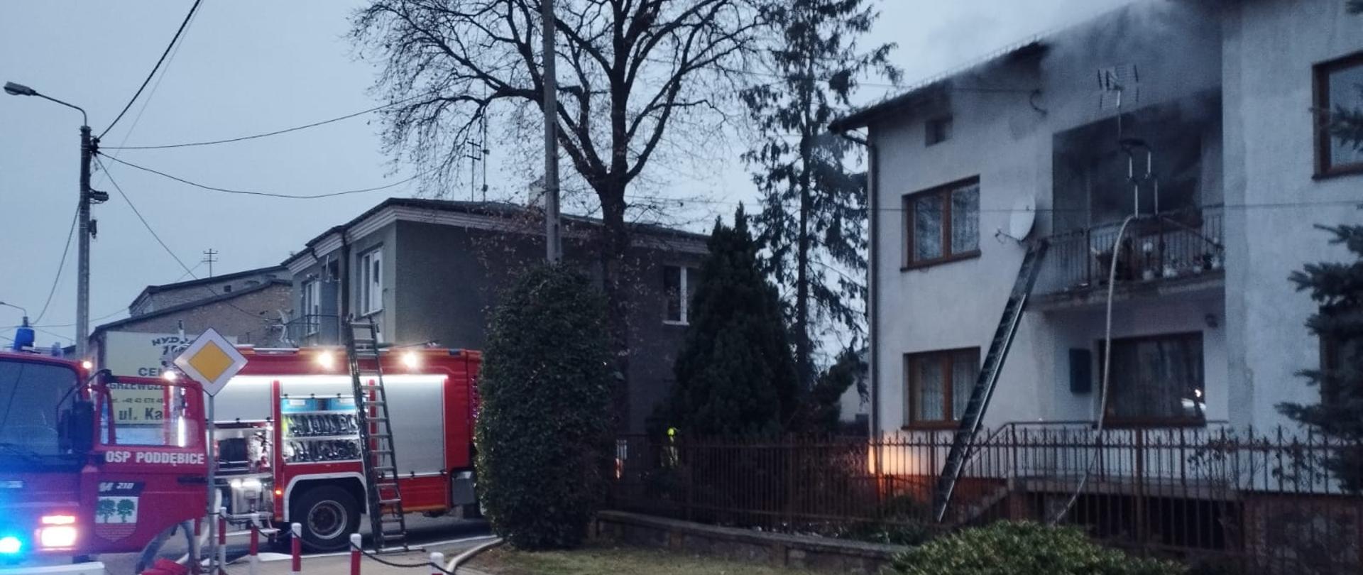 Piętrowy budynek. do balkonu który znajduje się z przodu budynku przystawiona jest drabina po której wchodzi strażak. Okno i drzwi balkonowy wyjęte z otworów wydostaje się dym. Obok budynku stoją dwa wozy strażackie 