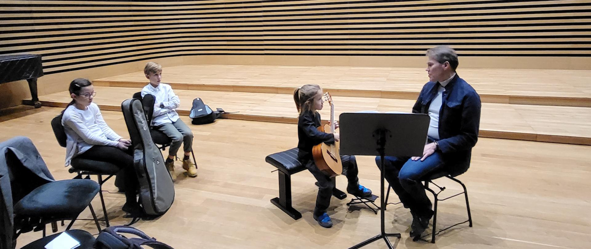 Dwóch uczniów i uczennica z prowadzącym na scenie sali koncertowej PSM