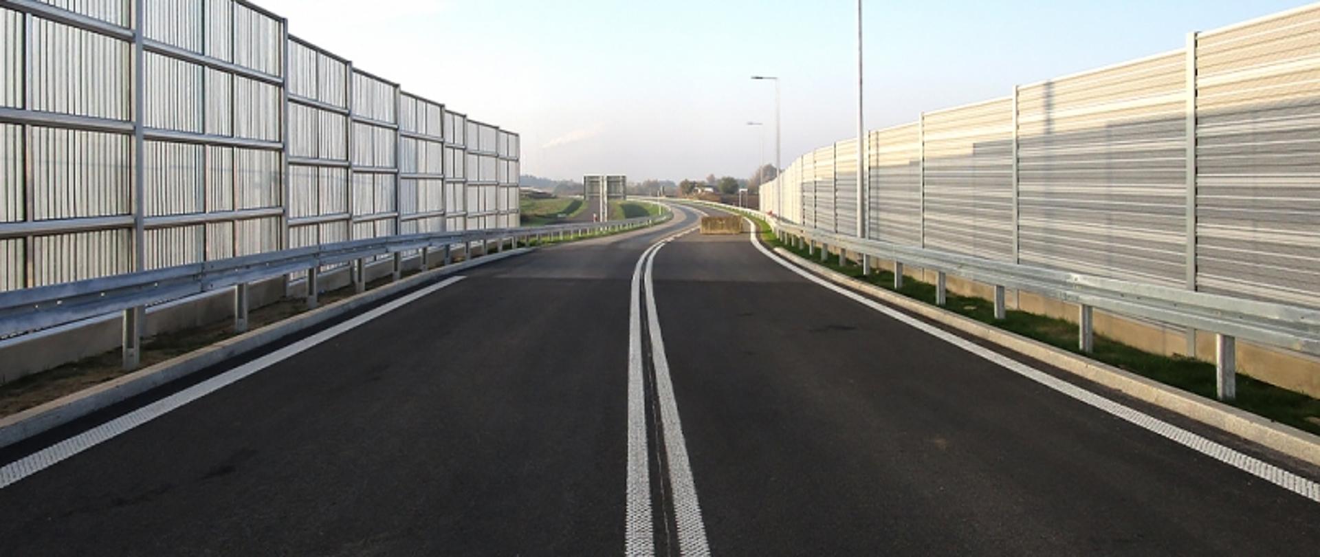 Dwupasmowa jezdnia z białymi liniami przerywanymi, metalowymi barierami ochronnymi i ekranami akustycznymi; w tle otwarty teren i lekkie zachmurzenie.