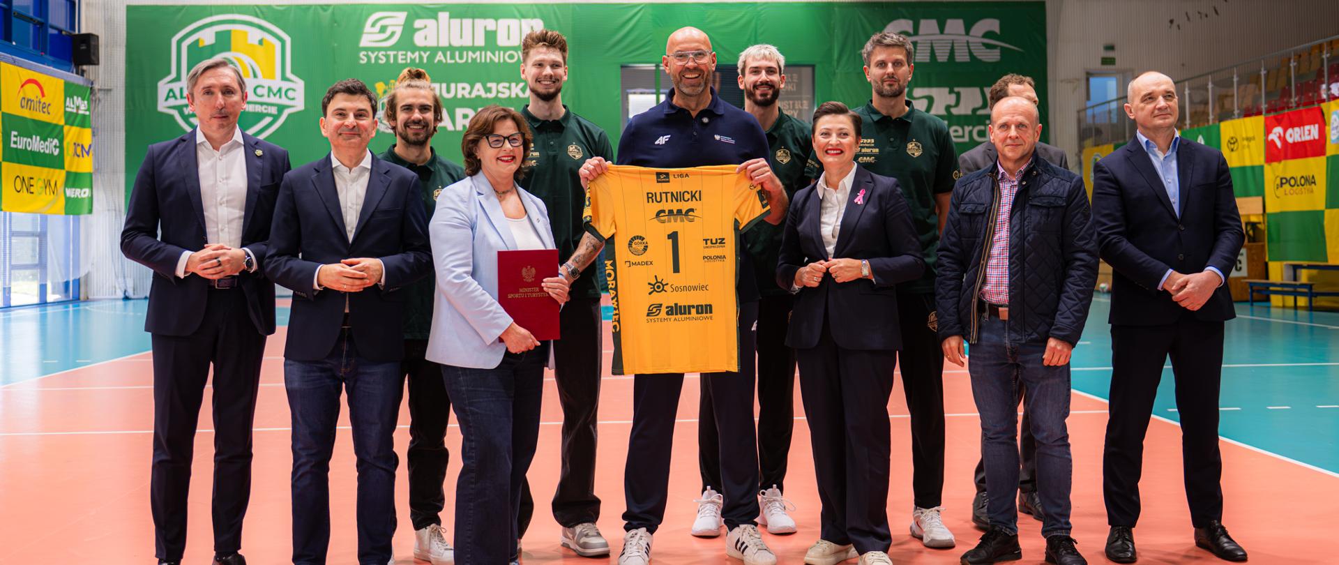 Zbiorowe zdjęcie uczestników konferencji prasowej w Zawierciu. 23 października 2026. Wśród nich reprezentanci Polski w siatkówce mężczyzn. Na środku stoi minister Sportu i Turystyki Jakub Rutnicki trzymający żółtą koszulkę Aluron CMC Warta Zawiercie z nazwiskiem Rutnicki. Zdjęcie zrobione w hali na boisku.