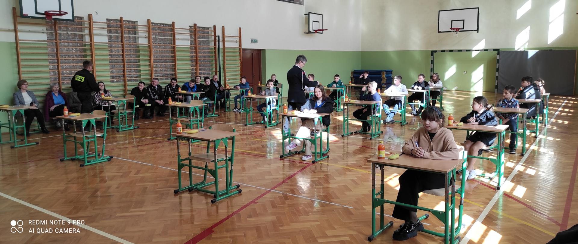 Kolorowa fotografia wykonana w pomieszczeniu sali gimnastycznej w szkole. Na sali znajdują się uczniowie którzy przystępują do części teoretycznej turnieju wiedzy pożarniczej. Na ławce z boku znajdują się strażacy OSP w mundurach i nauczyciele. Jeden ze strażaków w mundurze galowym rozdaje kartki z testami.