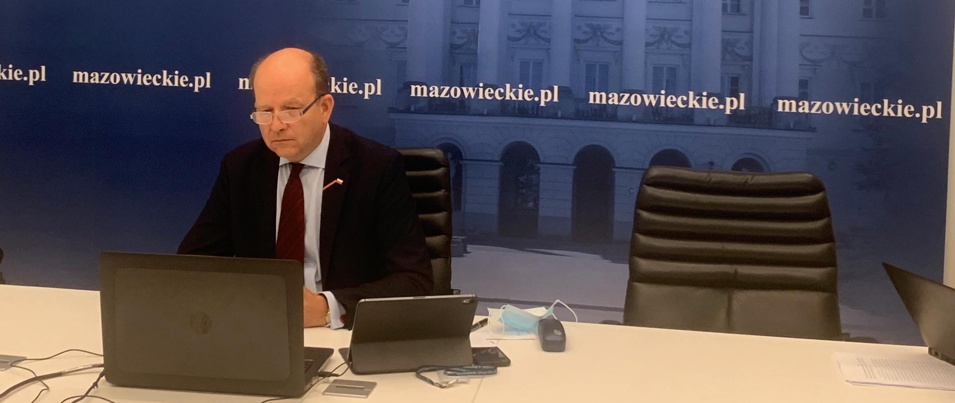 Na fotografii znajduje się ubrany w ciemną marynarkę, jasną koszulę i czerwony krawat mężczyzna. Mężczyzna ten siedzi za stołem, mając przed sobą laptopa. W tle znajduje się ścianka wizerunkowa z napisami "mazowieckie.pl" oraz logo Wojewody Mazowieckiego w lewym górnym rogu. Na ściance tej znajduje się także gmach siedziby Mazowieckiego Urzędu Wojewódzkiego.