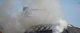 Na zdjęciu widoczni strażacy w ubraniach specjalnych koloru piaskowego w białych hełmach podający prąd wody w natarciu z podnośnika hydraulicznego na dach podczas pożaru obiektu handlowego przy ulicy Bieckiej w Gorlicach