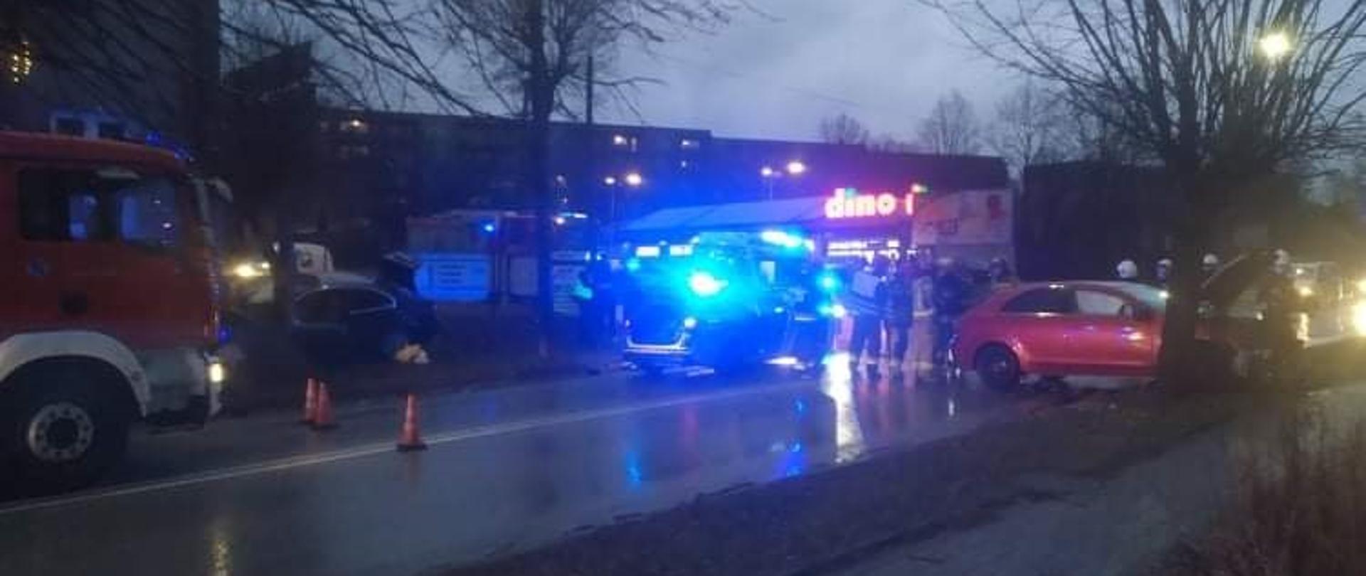 Zdjęcie obrazuje wypadek dwóch samochodów osobowych: Mitsubishi oraz BMW. Samochód BMW w wyniku zderzenia wypadł z drogi. Znajduje się on pomiędzy drzewem a budynkiem mieszkalnym. Ma uszkodzony cały przód. Samochód Mitsubishi znajduje się na pasie ruchu, ma rozbity cały przód. Miejsce wypadku zostało zabezpieczone przez strażaków pachołkami ostrzegawczymi.